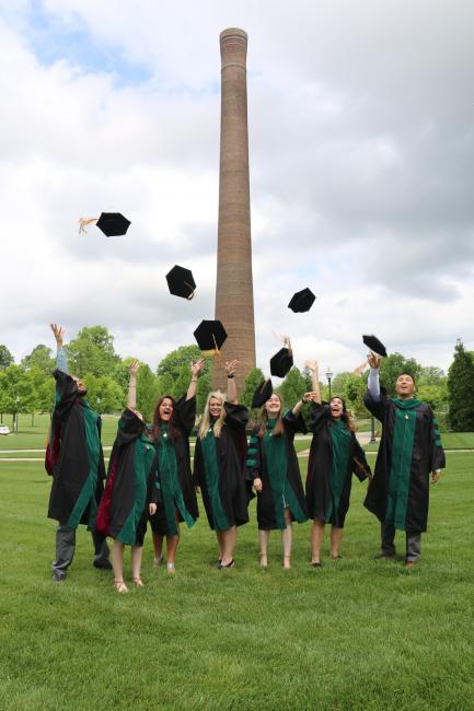 VCOM Carolinas graduates throw their caps into the air in front of the historic campus smoke stack
