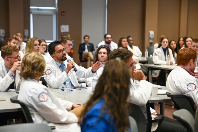 VCOM-Virginia CORE Symposium students in white coats with one person holding a microphone speaking