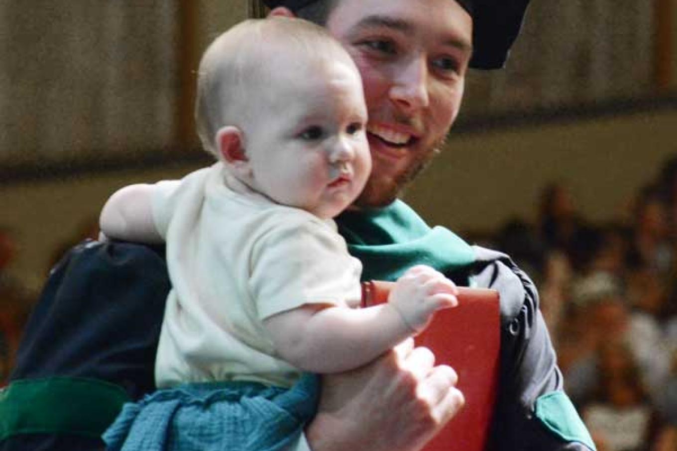 VCOM graduate in regalia holding his infant child