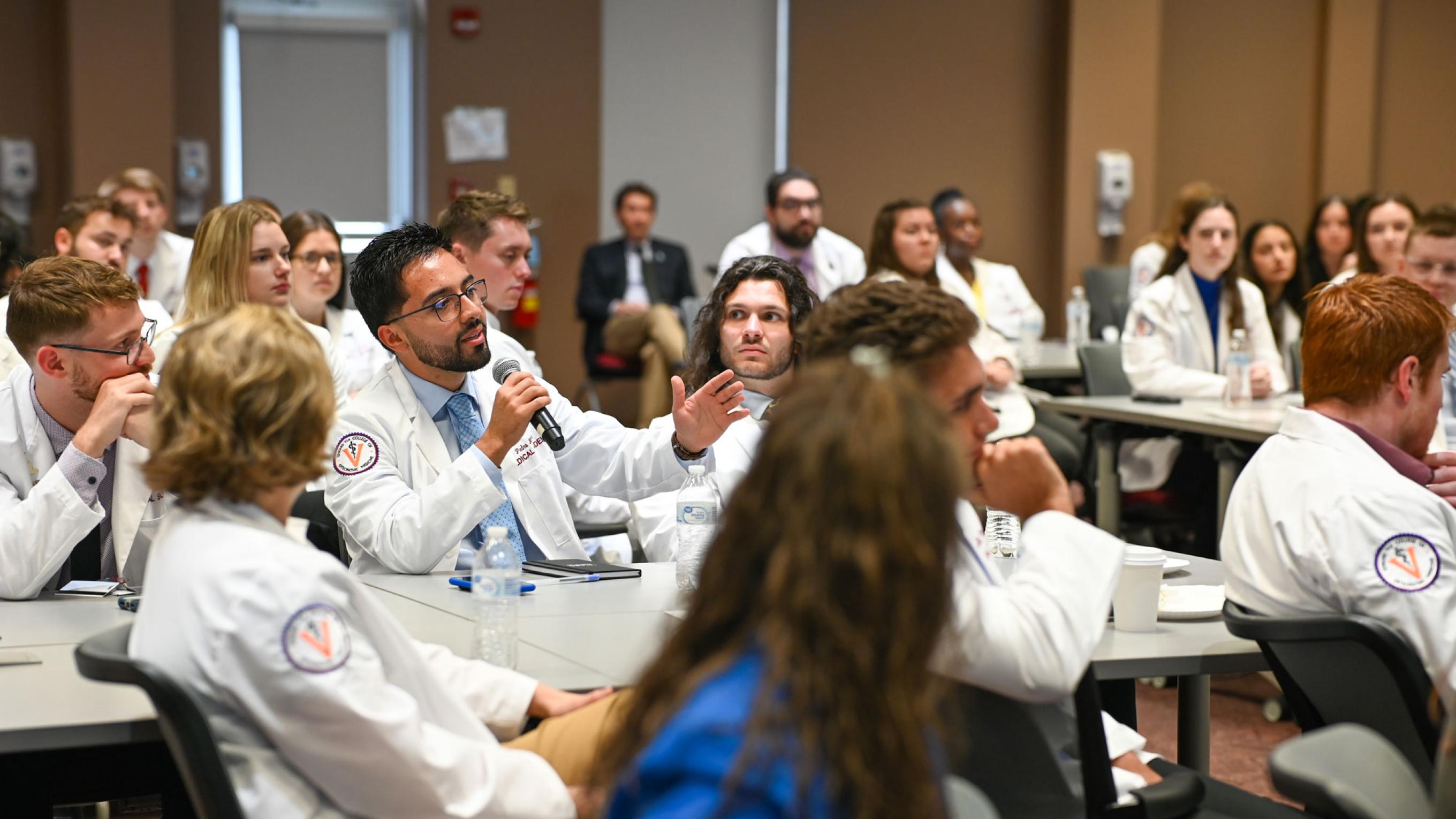 VCOM-Virginia CORE Symposium students in white coats with one person holding a microphone speaking 