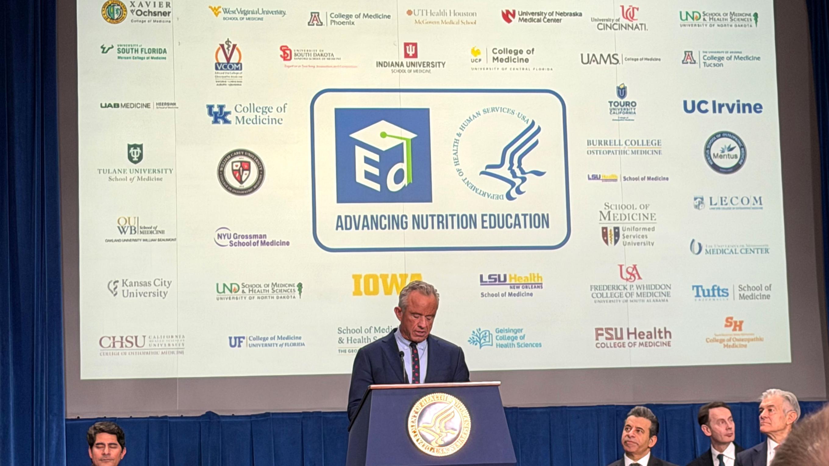 RFK Jr speaking in front of a screen of medical school logos