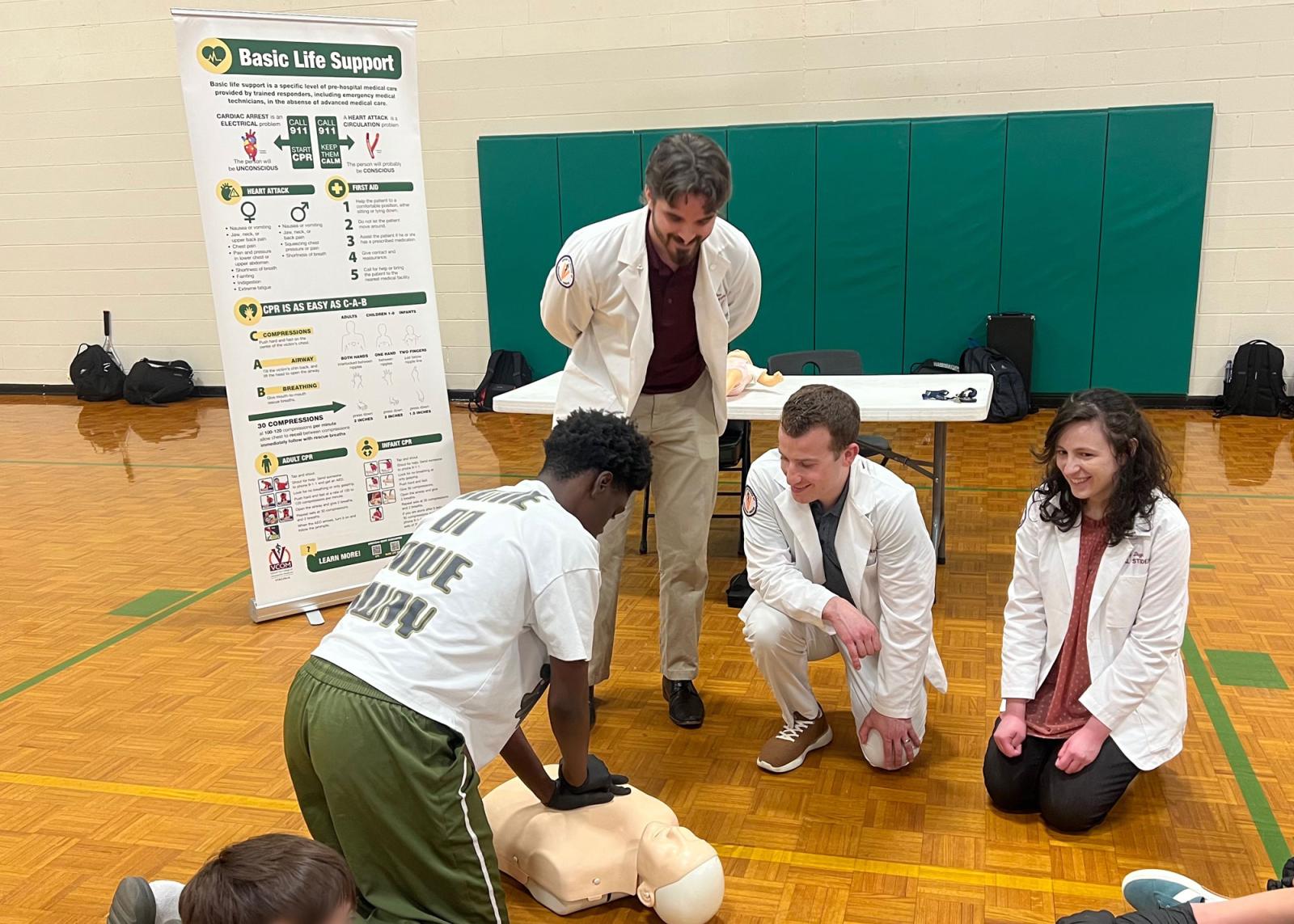 Students in white coats watching a high school student perform CPR on a manikin. 
