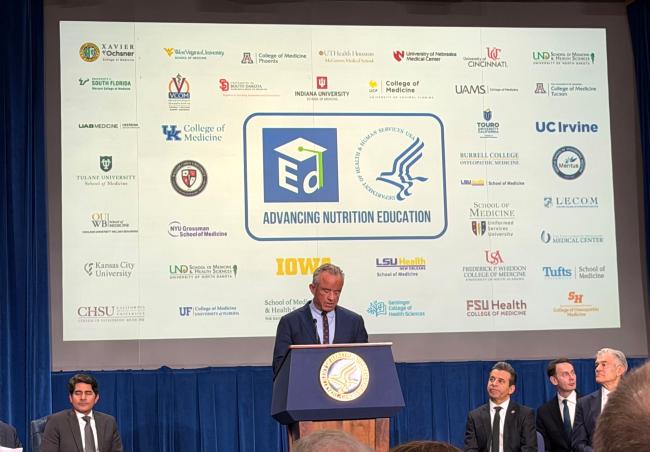 RFK Jr speaking in front of a screen of medical school logos