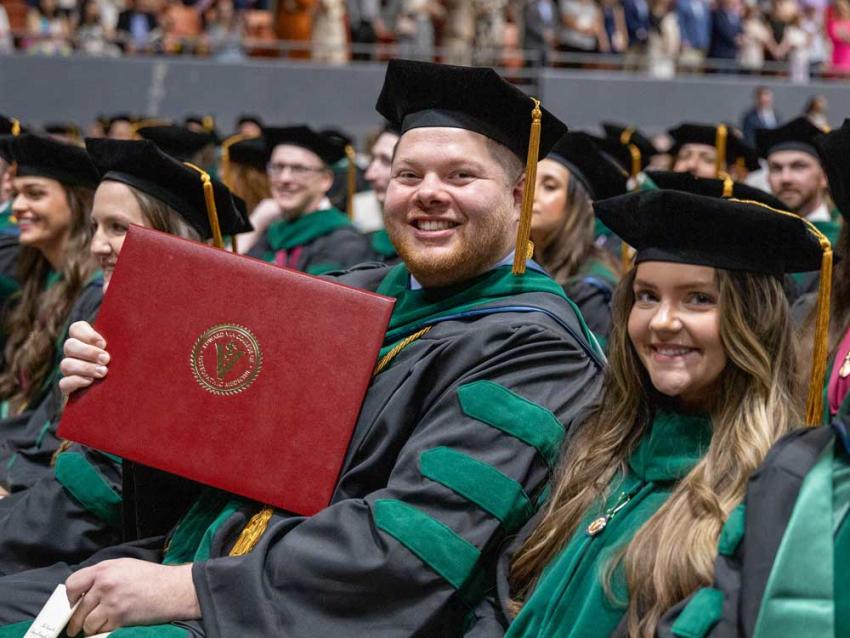 VCOM-Louisiana graduates in regalia holding diploma and smiling